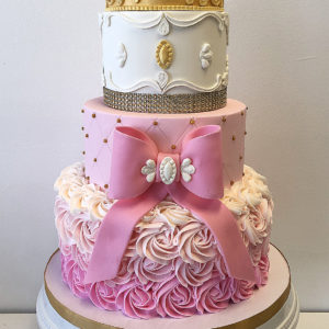 Three tiered birthday cake with a fondant bow and buttercream ombre rosettes. On top is a fondant gold painted crown.