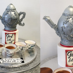 Small fondant cake with teapot on top carved of cake. The board has gumpaste teacups for each family member.