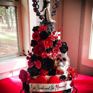 Three tiered fondant cake covered in black and red sugar roses. On the side is a hand made and painted sugar skull and in the front a hand painted fondant banner. 